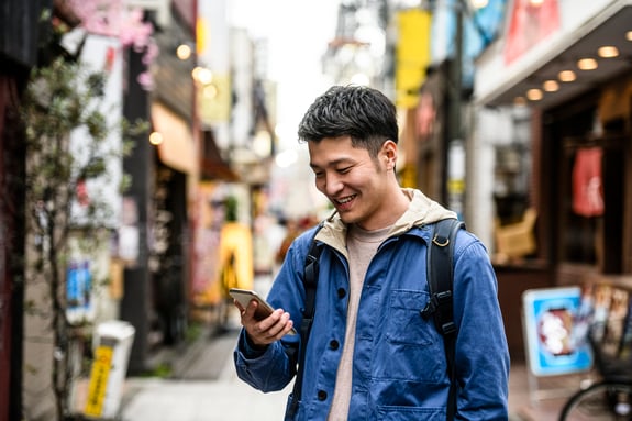An image of a man in a blue jacket, looking at his phone in the middle of a market. The man has dark hair and is smiling.